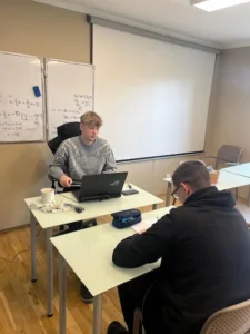 Lektor v šedé mikině individuálně pomáhá studentovi u pracovního stolu.